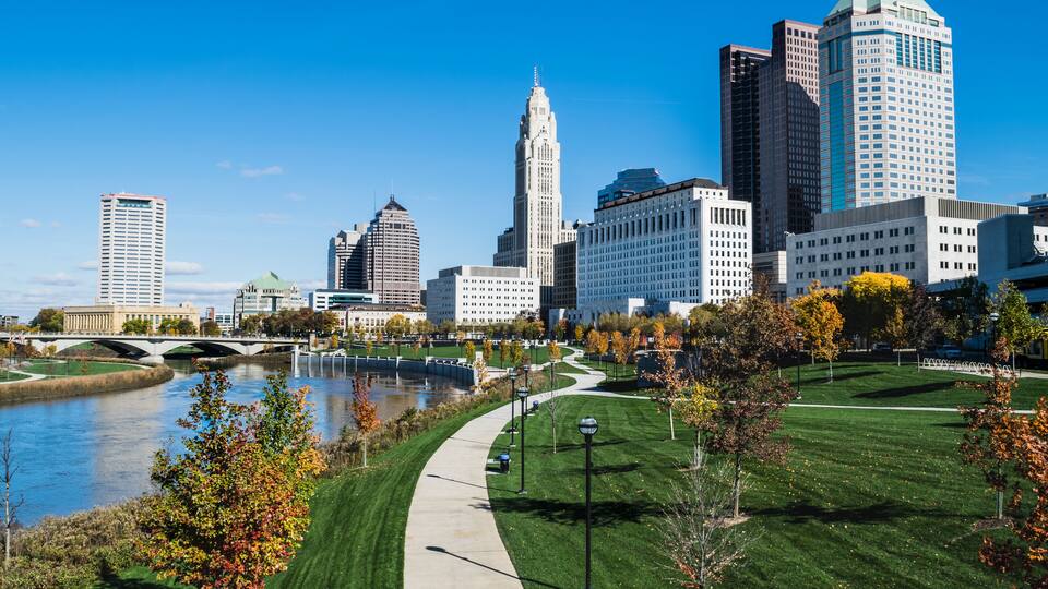 A cityscape with a river, a bridge, and a park with autumn trees.