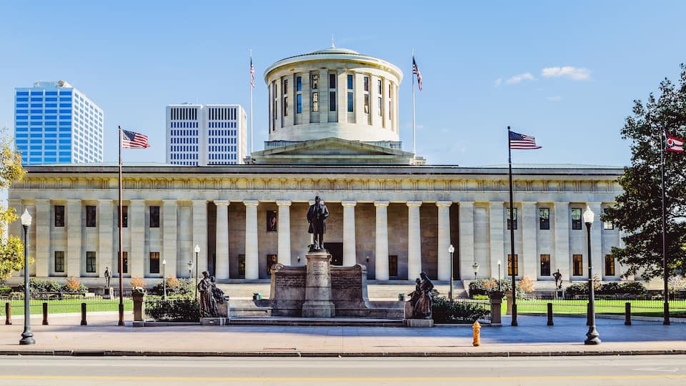 A neoclassical building with a central dome, flanked by columns, and two American flags.