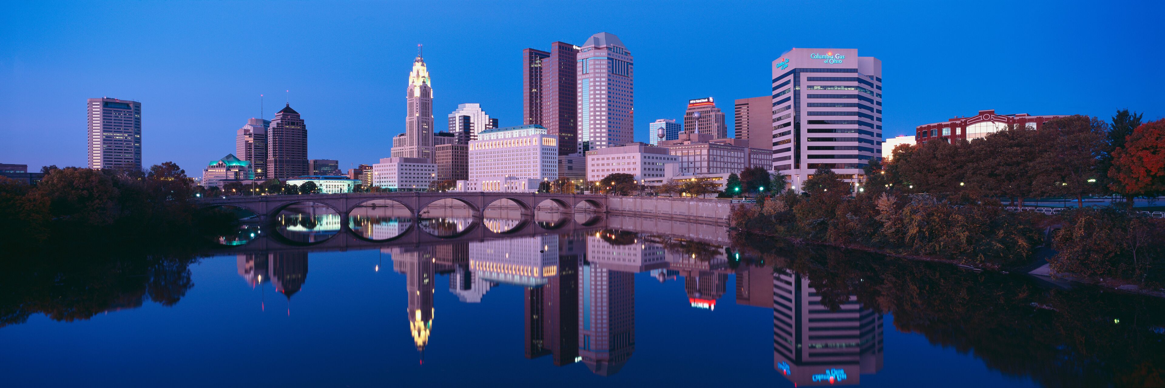 A city skyline reflected in a calm body of water, with a bridge in the foreground and various high-rise buildings.