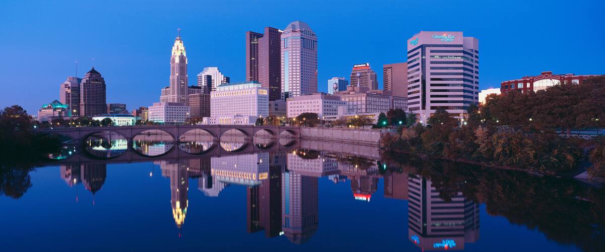 A city skyline reflected in a calm body of water, with a bridge in the foreground and various high-rise buildings.