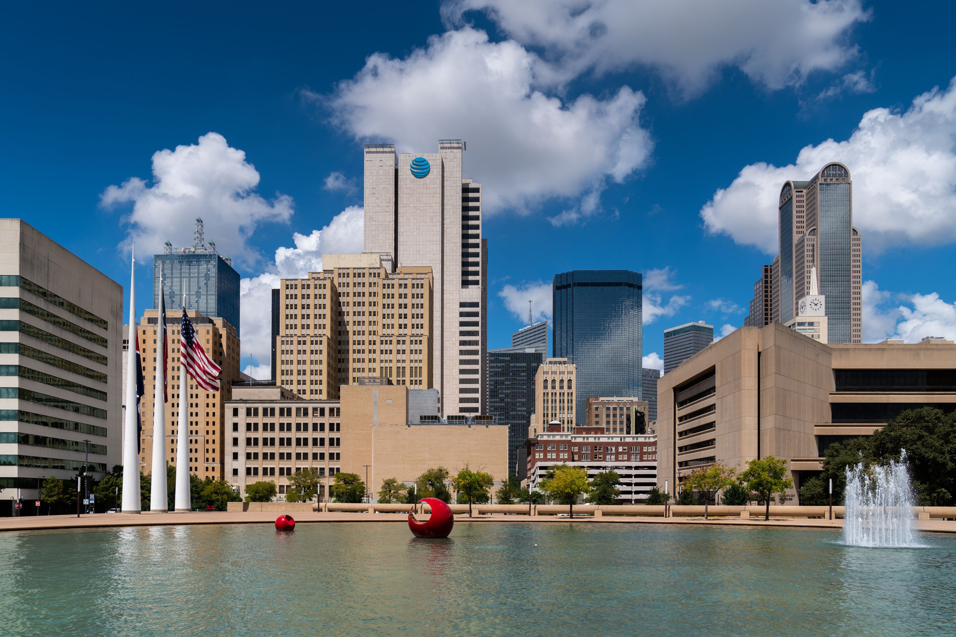 A city skyline with tall buildings, a body of water with a red sphere and a fountain, and American flags.
