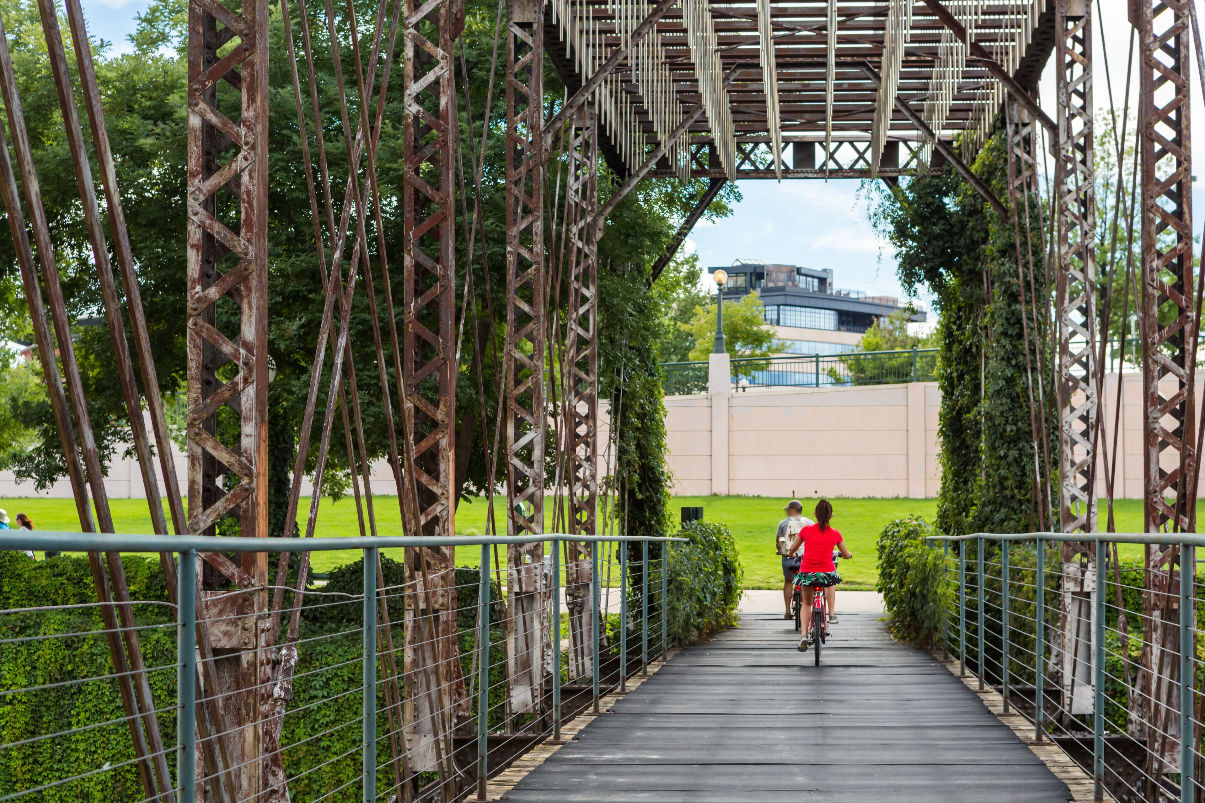 A pedestrian bridge with metal railings and a pathway, surrounded by greenery and a building in the background.