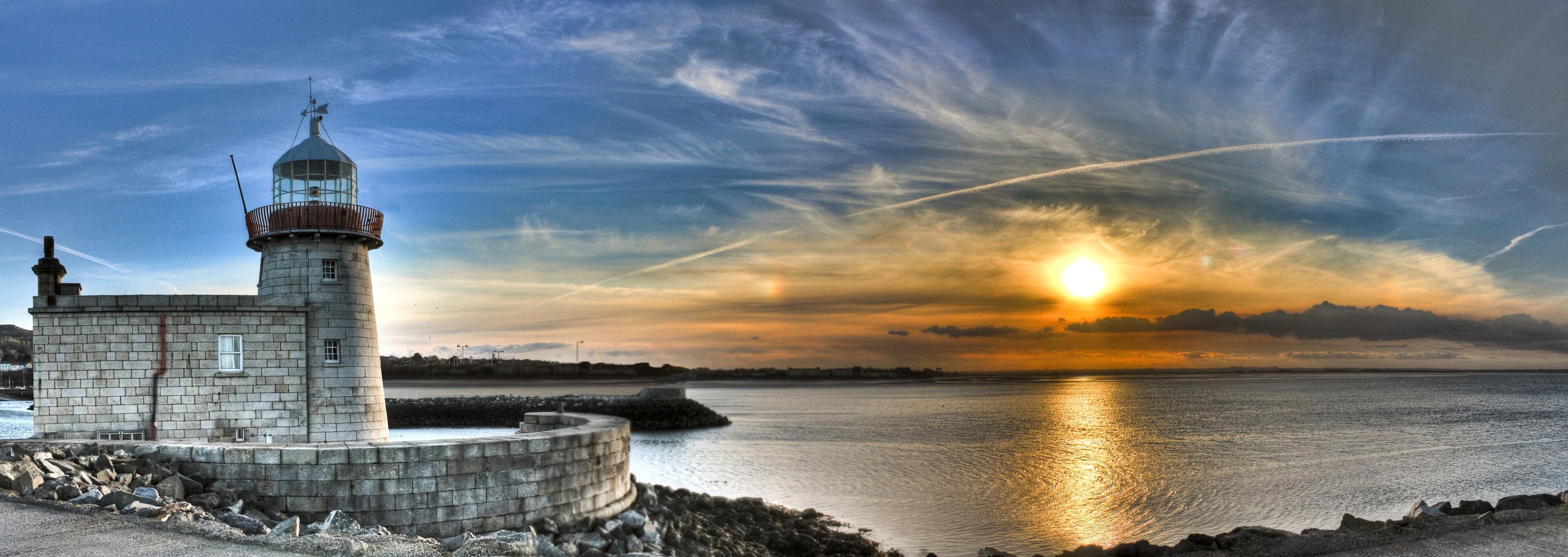 A lighthouse by a calm sea at sunset with a clear sky.