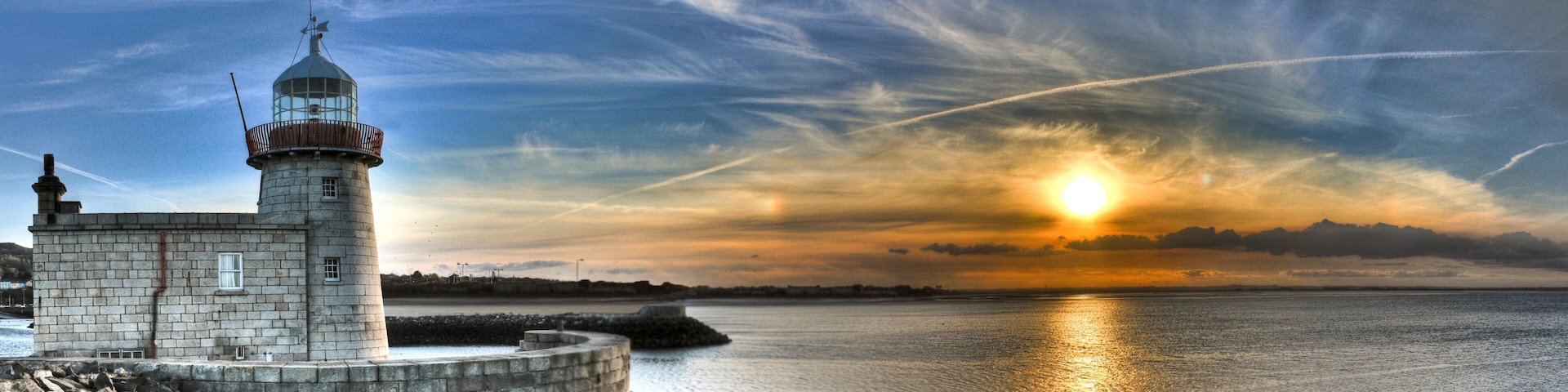 Un faro frente a un mar tranquilo al atardecer, con cielo despejado.