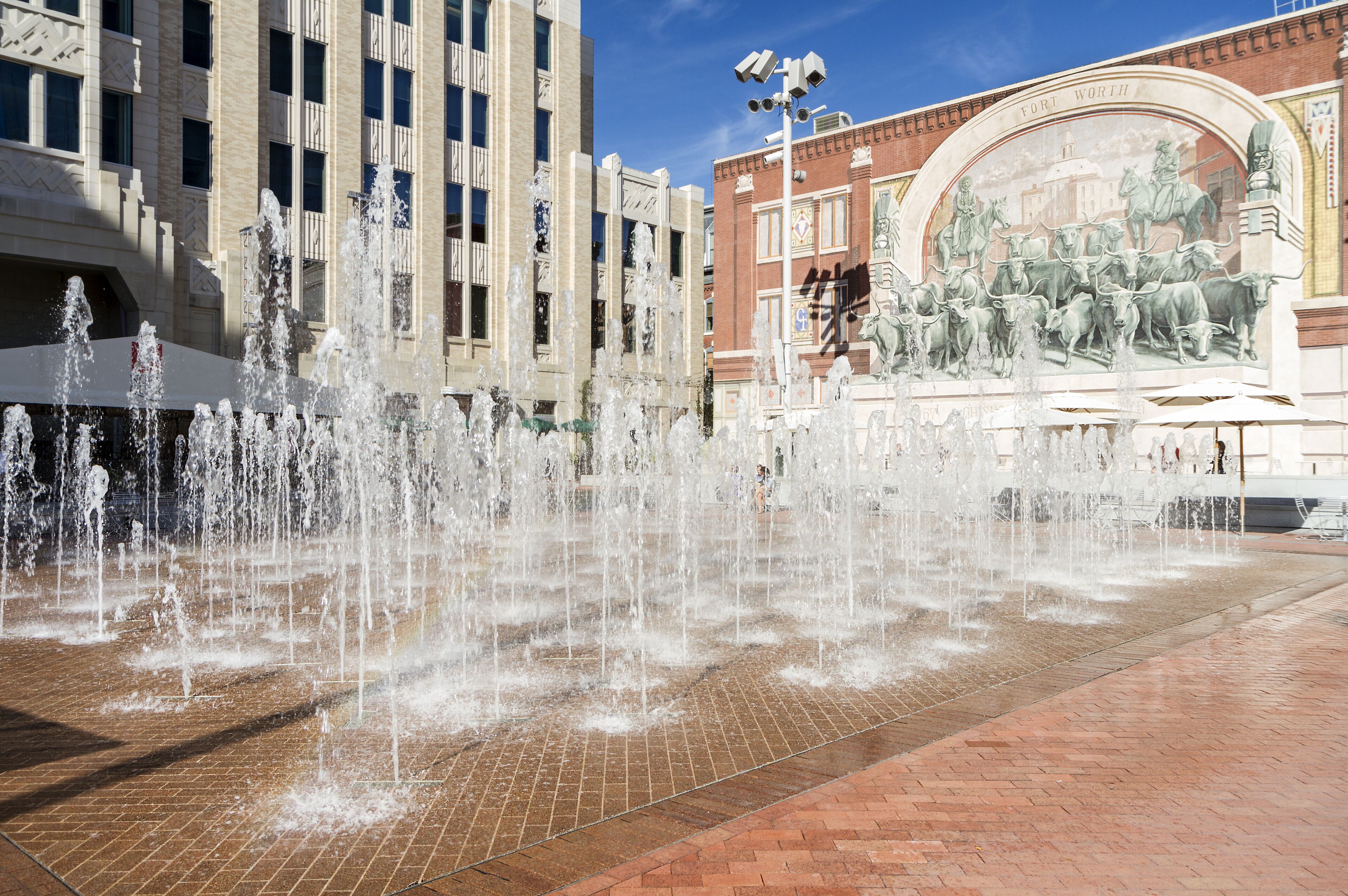 A fountain with water jets in a plaza, surrounded by buildings and a mural.