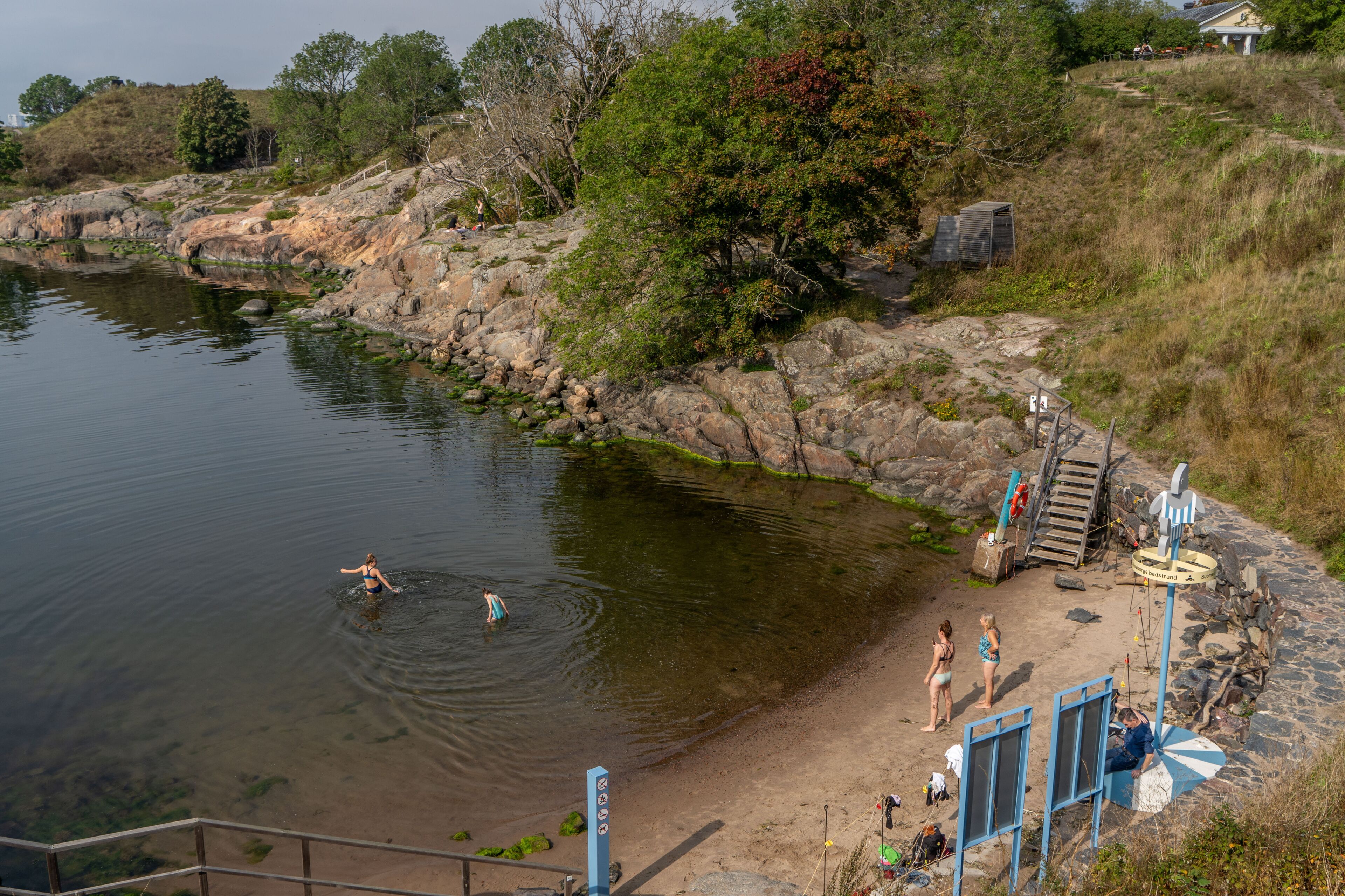 A river with people swimming, a sandy beach, and a rocky area with trees.