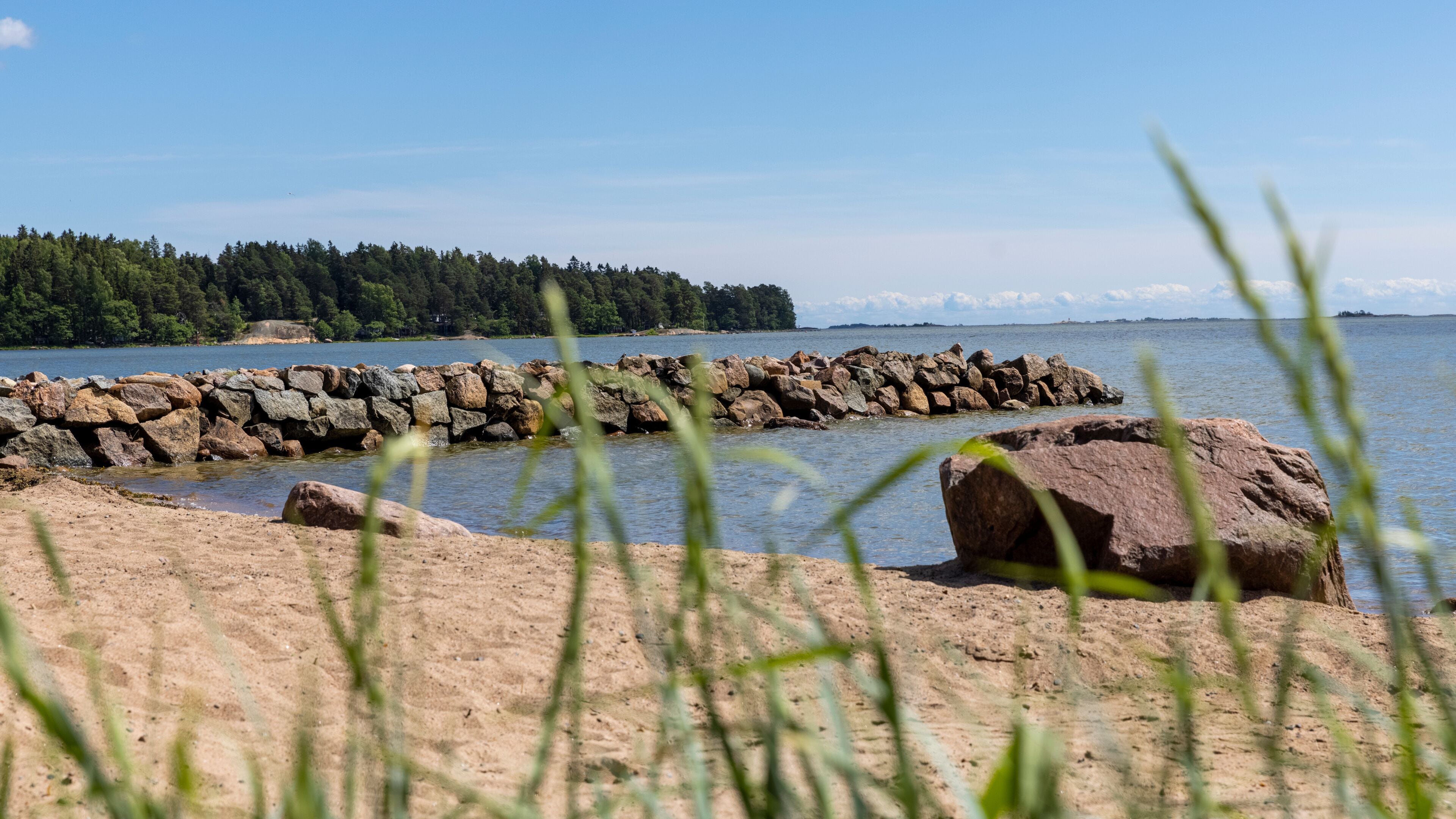 A rocky shoreline with a stone breakwater extending into a calm lake, surrounded by dense forest and clear skies.