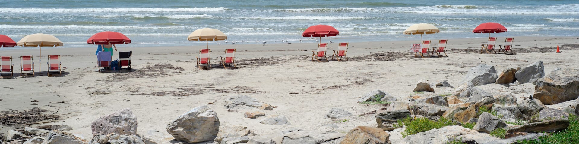 Panorama view quiet Galveston shoreline with large rock formations dominates foreground, empty beach umbrellas stand facing gentle waves, clear sky, peaceful solitude, ideal for coastal travel