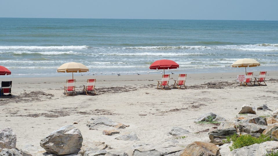 A beach with red and beige umbrellas, beach chairs, and large rocks in the foreground.