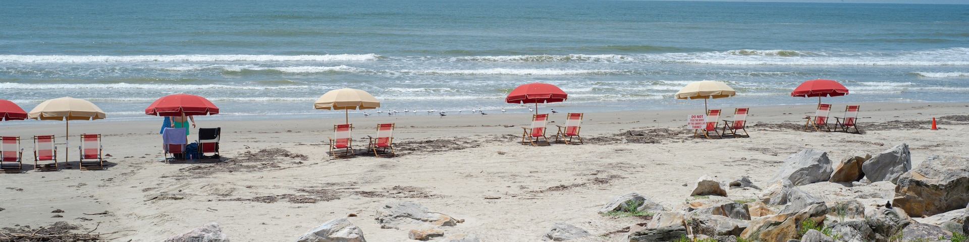 A beach with red and beige umbrellas, beach chairs, and large rocks in the foreground.