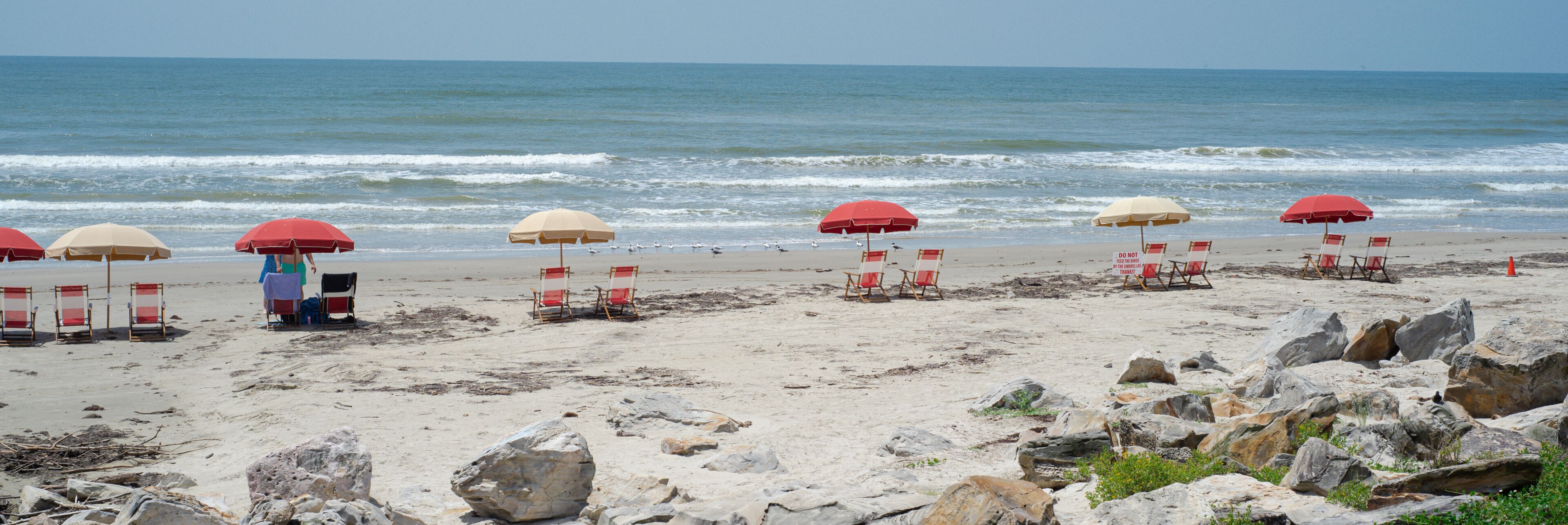 A beach with red and beige umbrellas, beach chairs, and large rocks in the foreground.