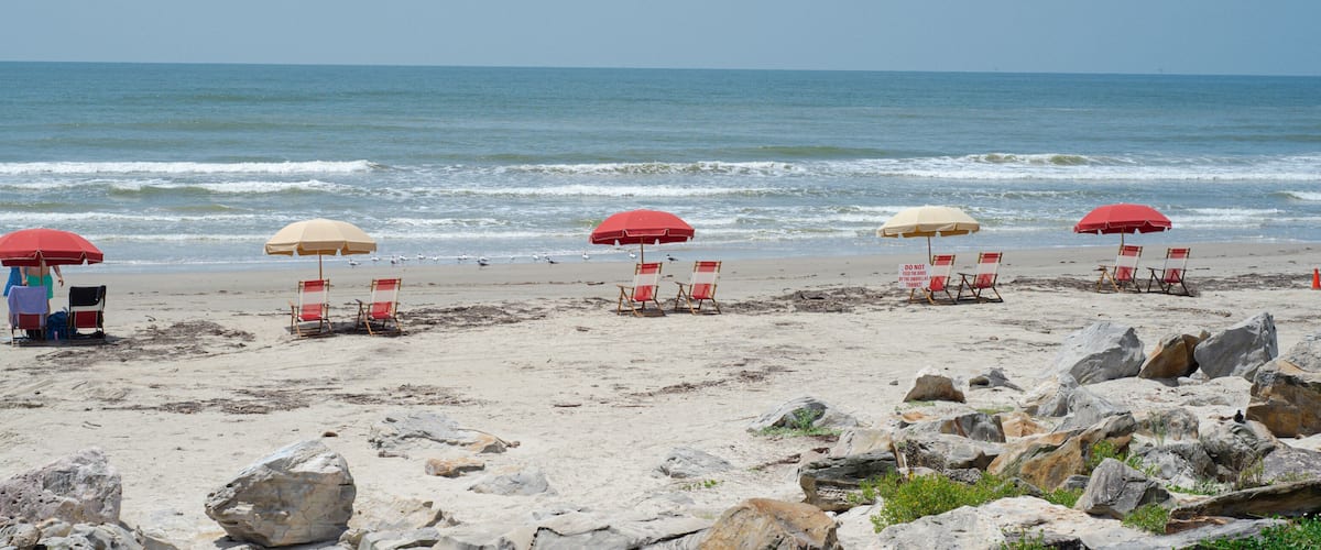 A beach with red and beige umbrellas, beach chairs, and large rocks in the foreground.