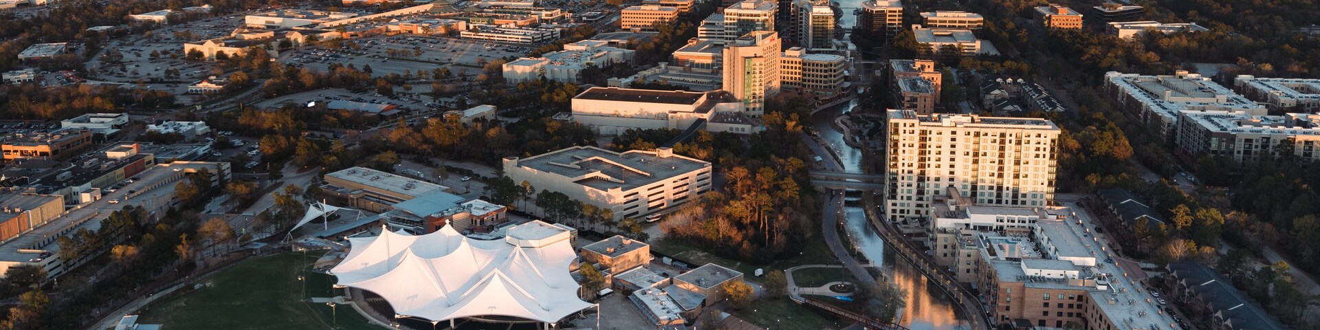 An aerial view of a cityscape with a prominent white-roofed stadium, a river, and surrounding buildings.