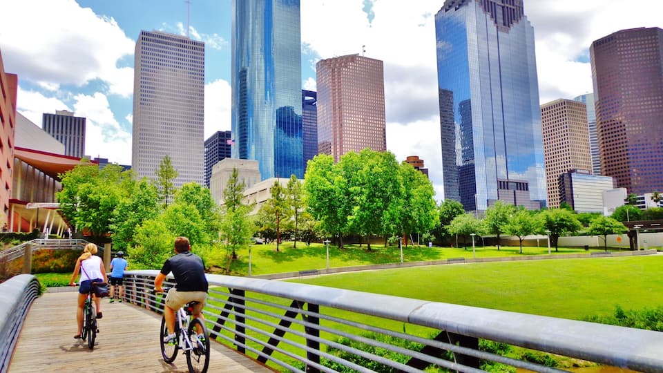 A cityscape with modern skyscrapers, a park with green trees, and a wooden bridge with cyclists.