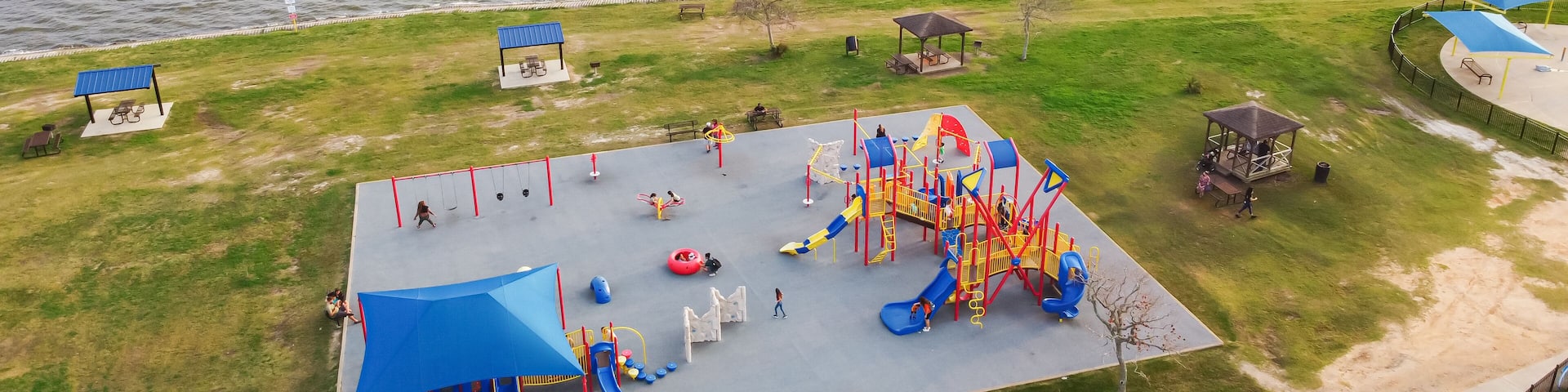 Panorama aerial view kids swing, climb, slide on colorful playground while parents supervise. Bright equipment and green lawns surround this lively outdoor scene, Clear Lake Park, Seabrook, Texas