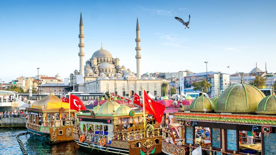 A traditional boat with ornate decorations and flags, docked near a mosque with two minarets and a dome, in a busy urban waterfront.