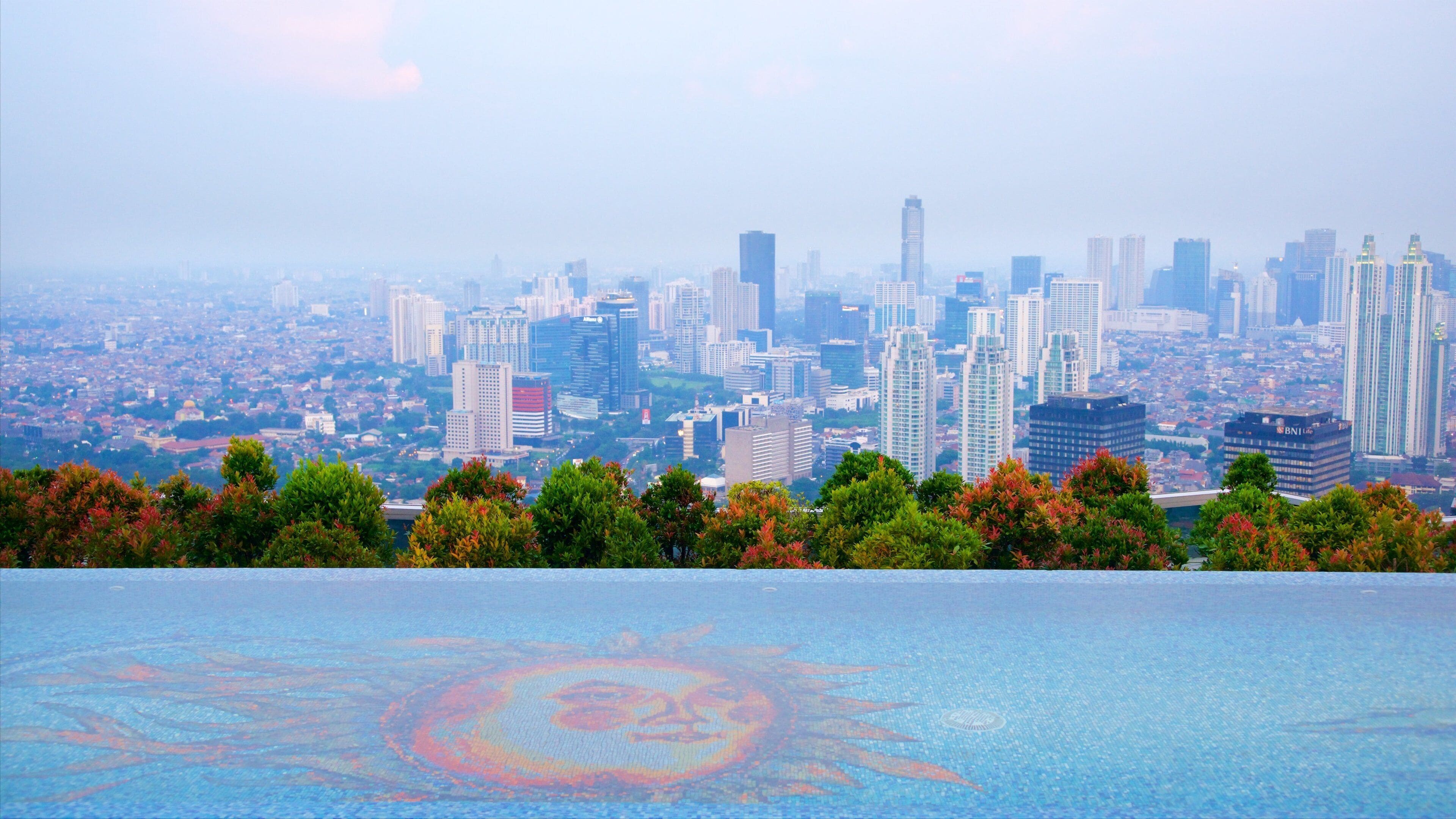 Paisagem urbana com edifícios altos e modernos e um design em forma de sol refletido em uma superfície.