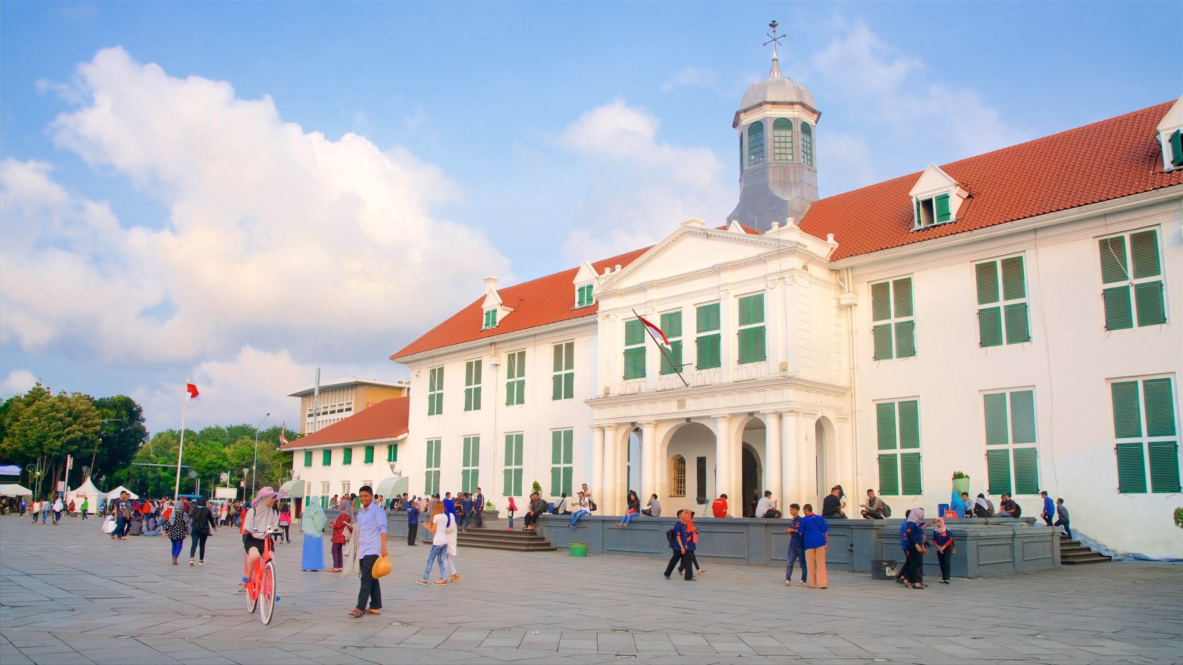 A historic building with a red-tiled roof and green shutters, surrounded by a public square with people walking and sitting.