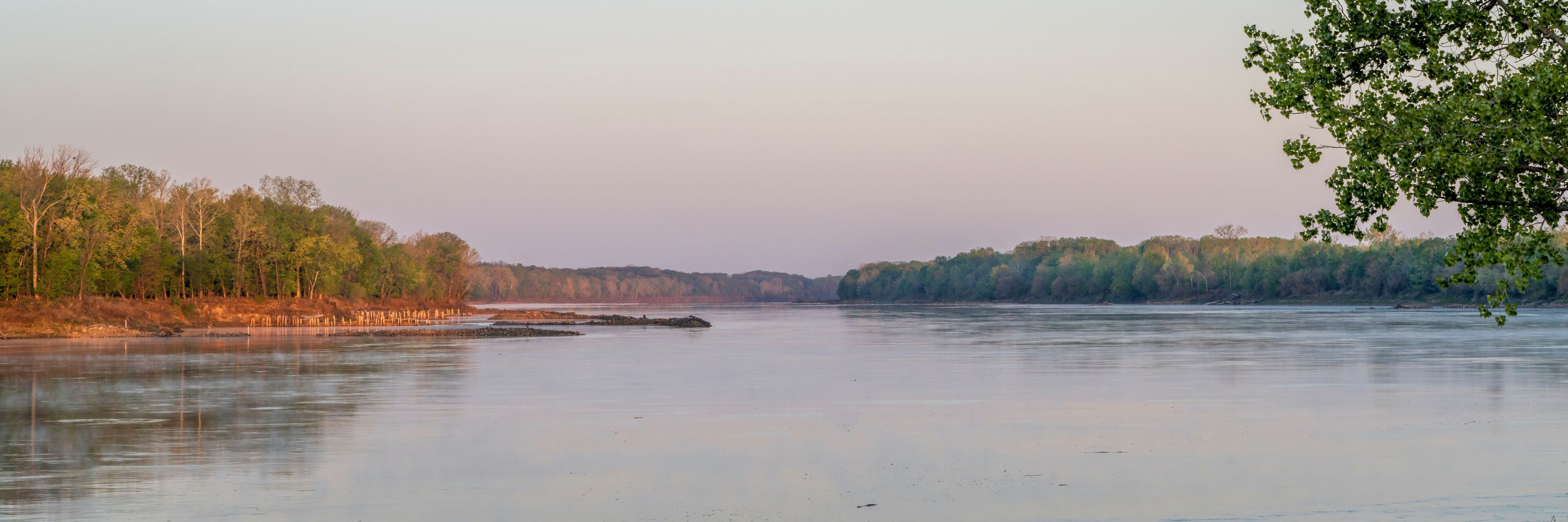 Un río tranquilo con árboles a ambos lados, cielo despejado y una costa lejana.