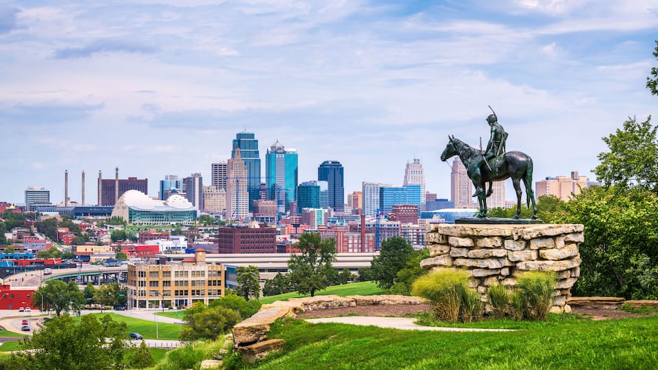 A city skyline with a statue of a mounted figure on a stone pedestal in the foreground.