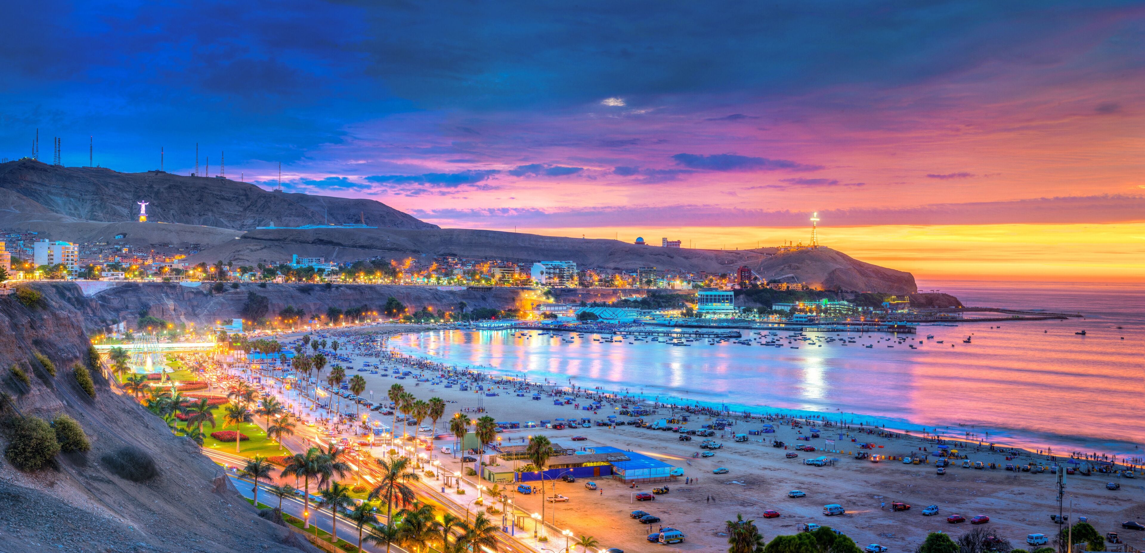 A coastal cityscape at sunset with a beach, palm trees, and illuminated buildings.