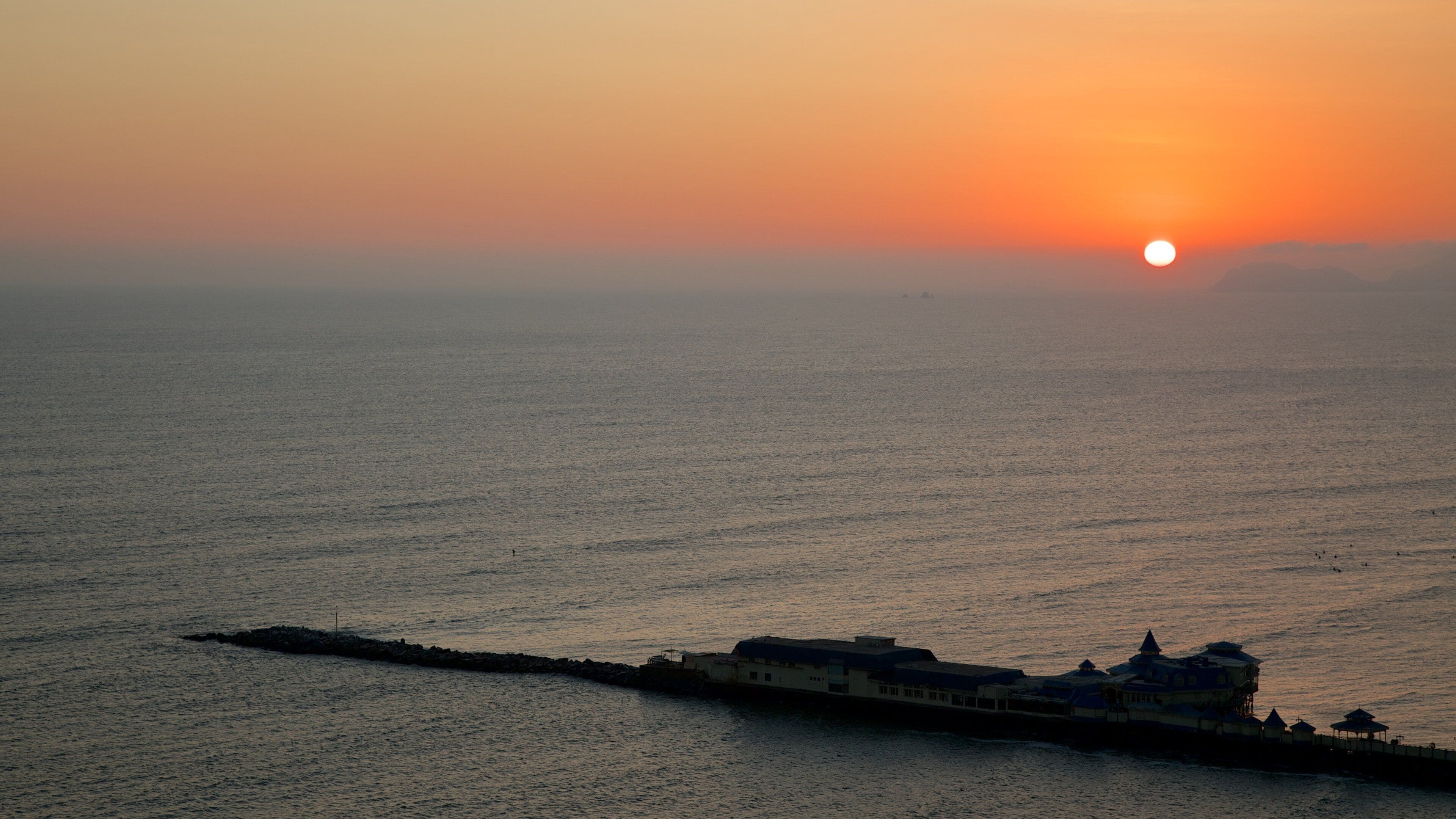 A sunset over a calm sea with a pier and buildings in the foreground.