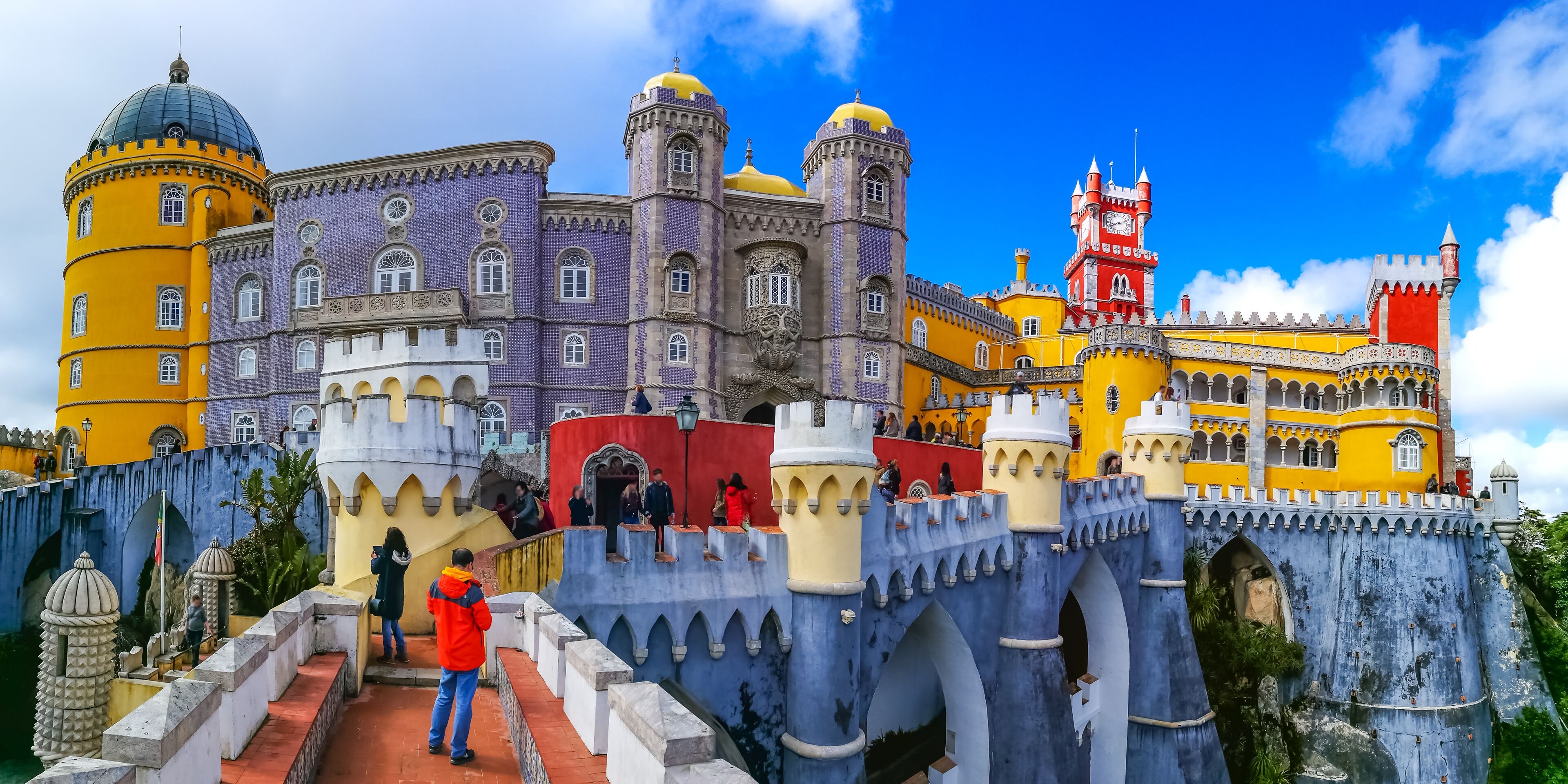 A colorful castle with yellow and purple walls, a red tower, and a blue bridge.