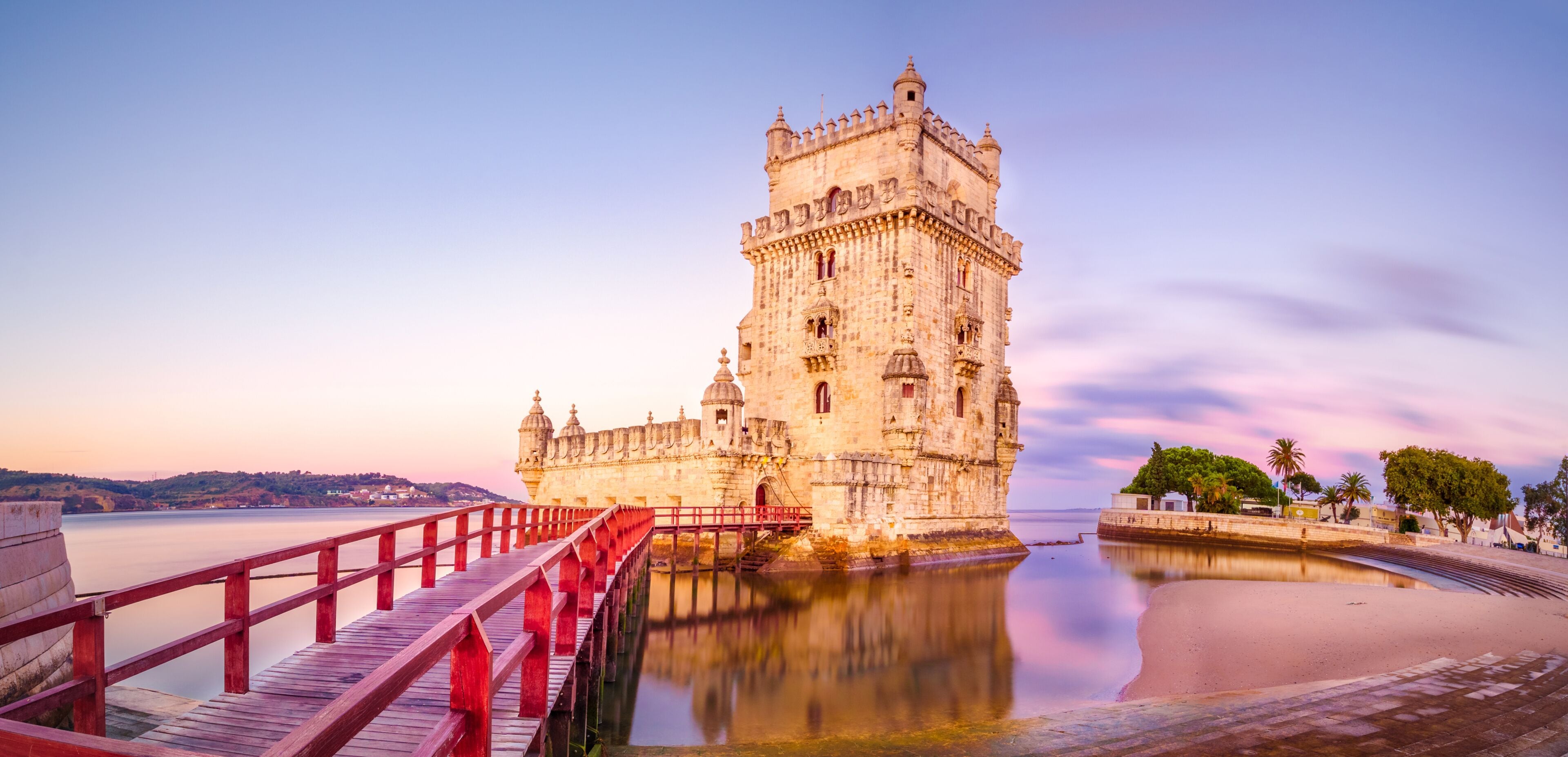A historic castle with a red bridge leading to it, situated by a calm body of water.