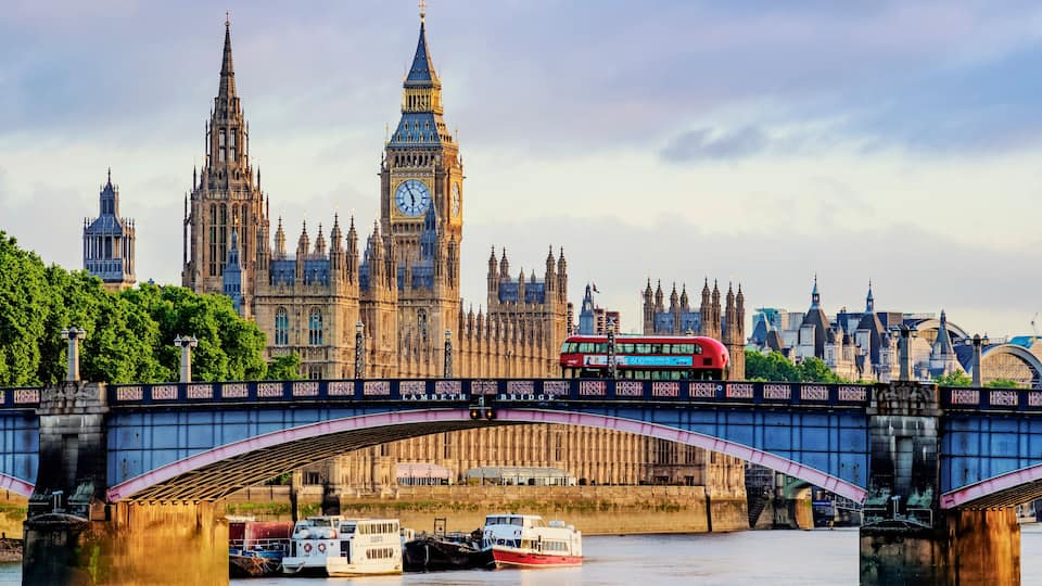 The Houses of Parliament and Big Ben in London, with a red double-decker bus crossing the Lambeth Bridge.