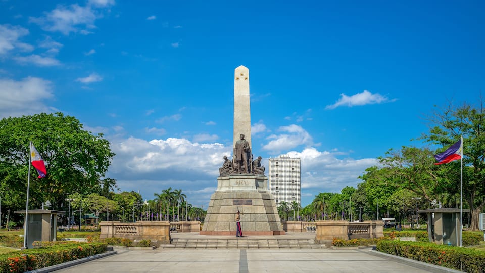 A monument with a statue, surrounded by trees and flags, under a clear blue sky.