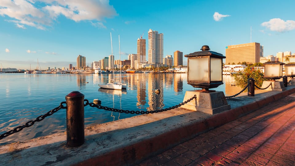 A waterfront cityscape with a clear sky, a chain-link fence, and a row of buildings along the water.
