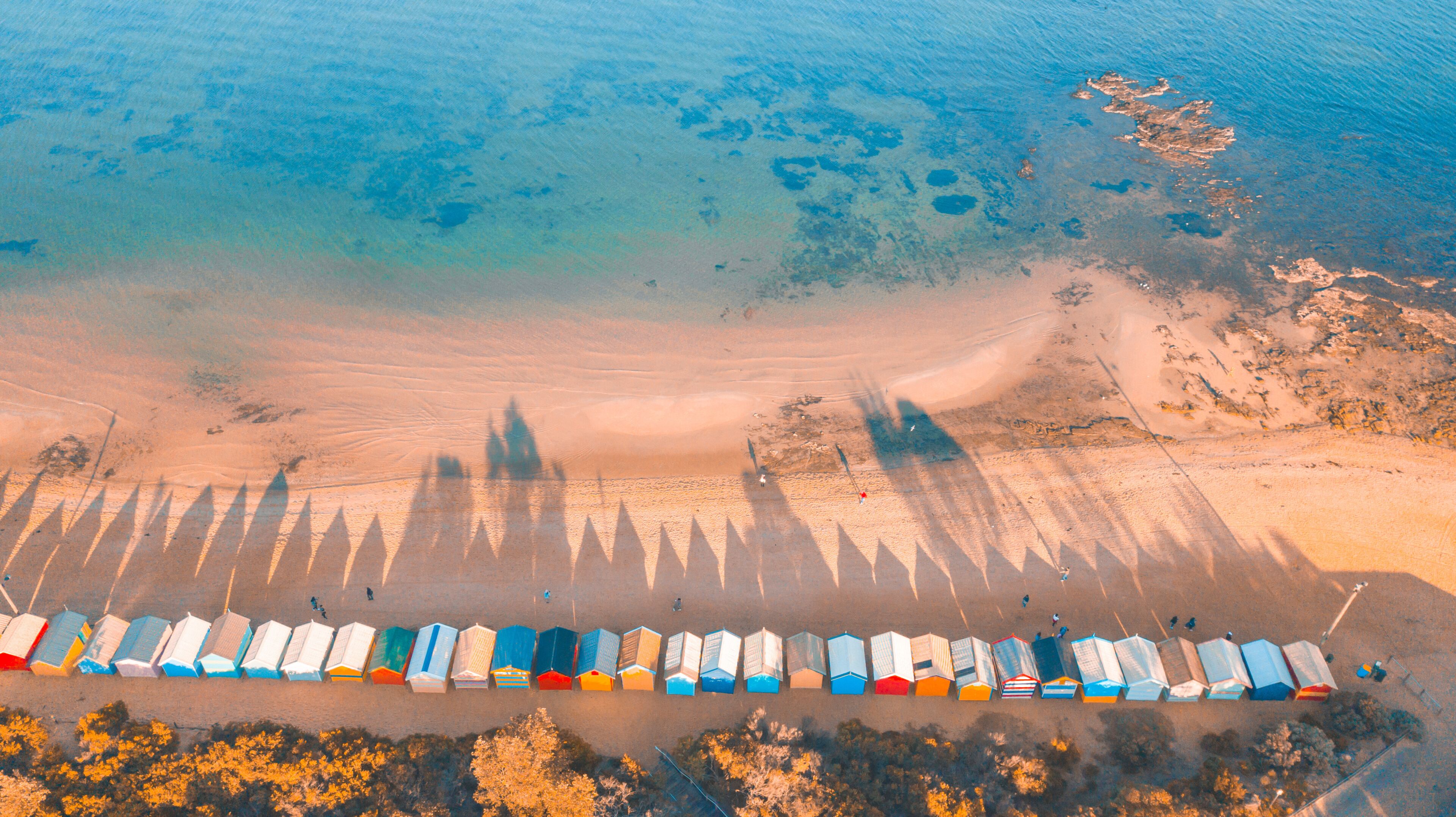 An aerial view of a beach with a row of colorful beach huts and shadows of people and objects.