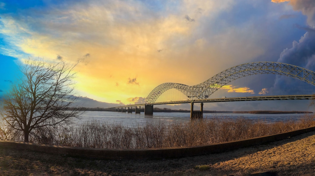 a stunning panoramic shot of the Memphis-Arkansas Bridge over the vast flowing waters of the Mississippi river with blue sky and powerful yellow clouds and dry brush on the banks of the river