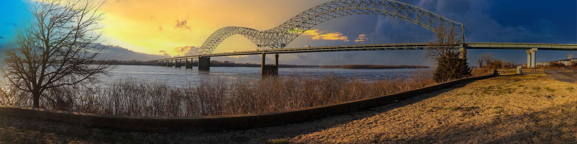 a stunning panoramic shot of the Memphis-Arkansas Bridge over the vast flowing waters of the Mississippi river with blue sky and powerful yellow clouds and dry brush on the banks of the river