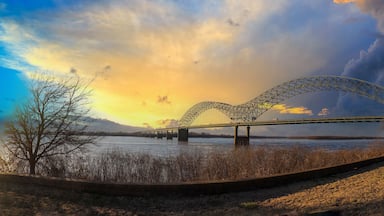 a stunning panoramic shot of the Memphis-Arkansas Bridge over the vast flowing waters of the Mississippi river with blue sky and powerful yellow clouds and dry brush on the banks of the river