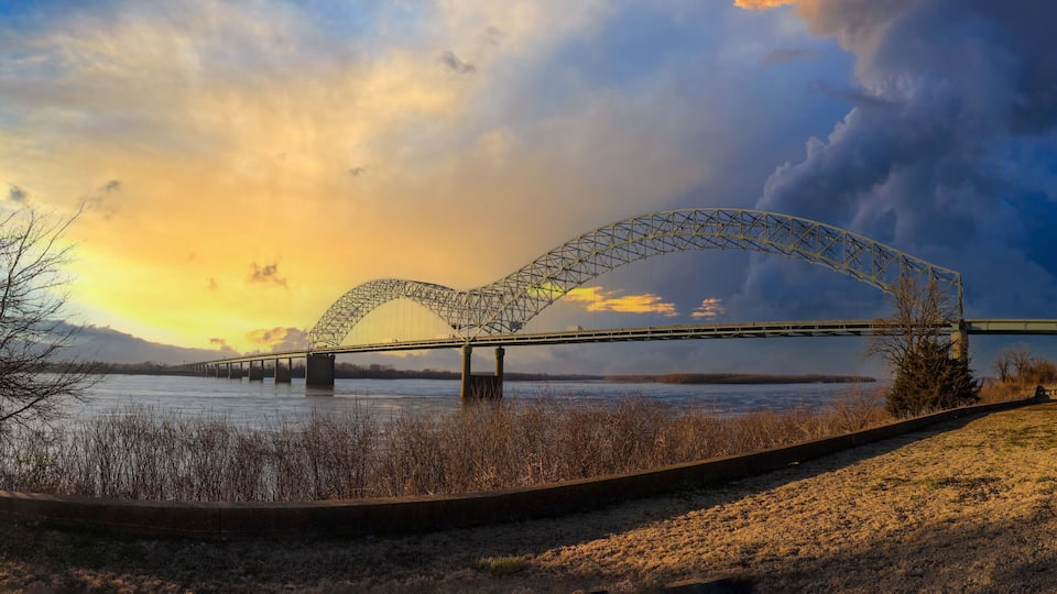 A bridge with a heart-shaped arch over a river, surrounded by trees and a cloudy sky.