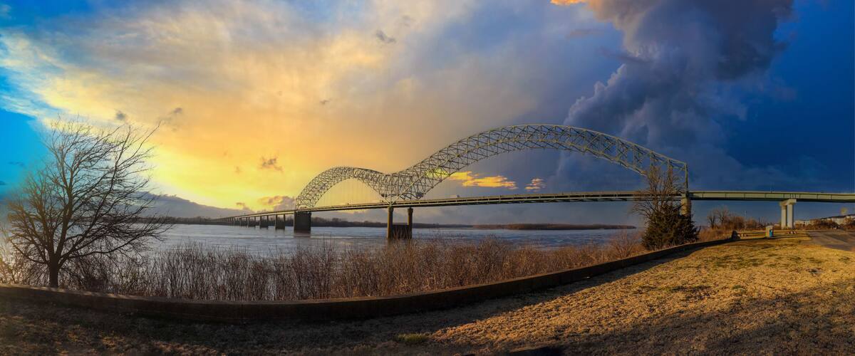 A bridge with a heart-shaped arch over a river, surrounded by trees and a cloudy sky.