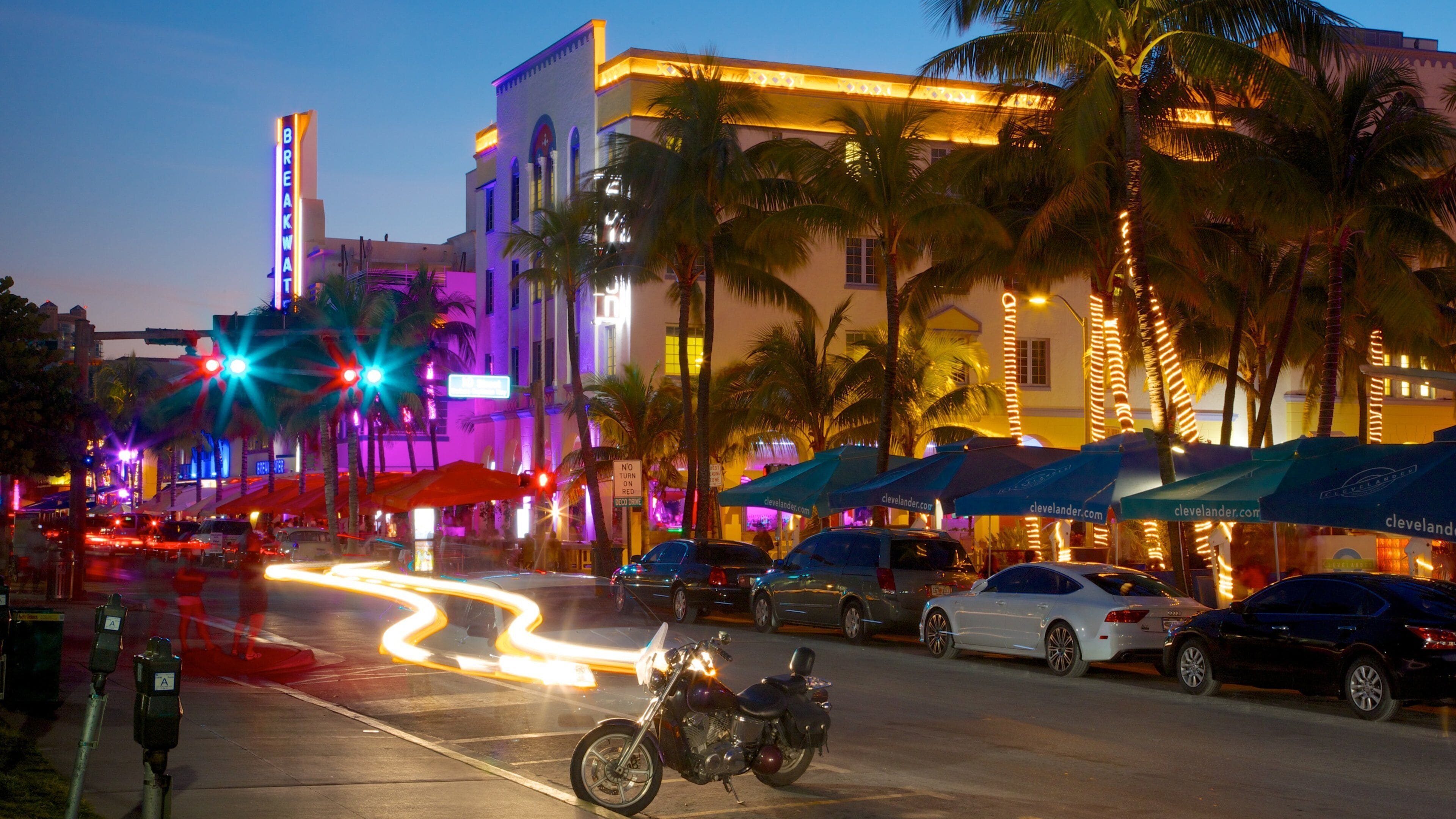 A busy street at dusk with palm trees, illuminated buildings, and parked cars.