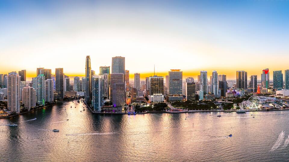 A city skyline at sunset with high-rise buildings, a waterfront, and a Ferris wheel.
