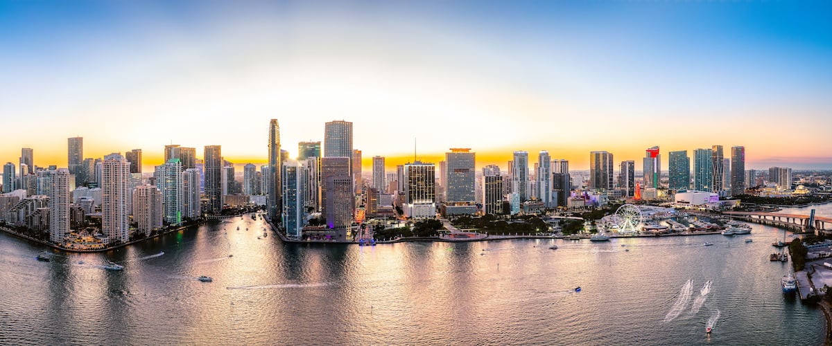 A city skyline at sunset with high-rise buildings, a waterfront, and a Ferris wheel.