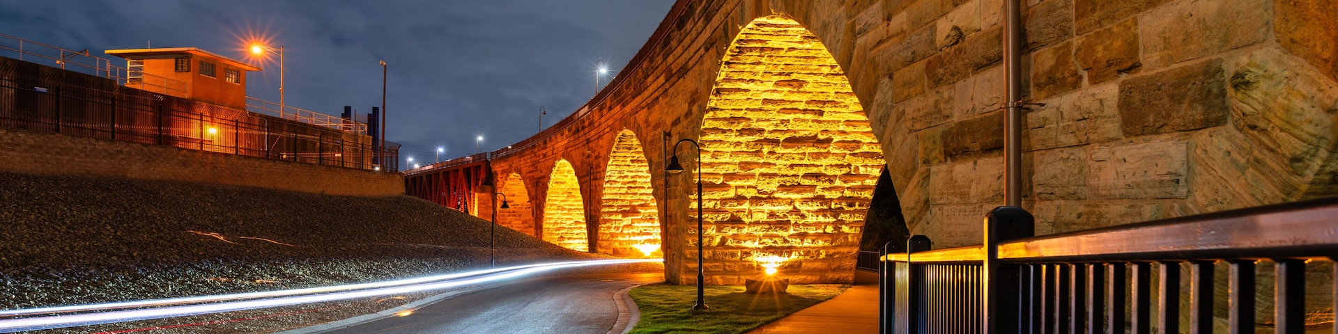 Uma ponte de pedra à noite com arcos iluminados e uma estrada com rastros de luz.