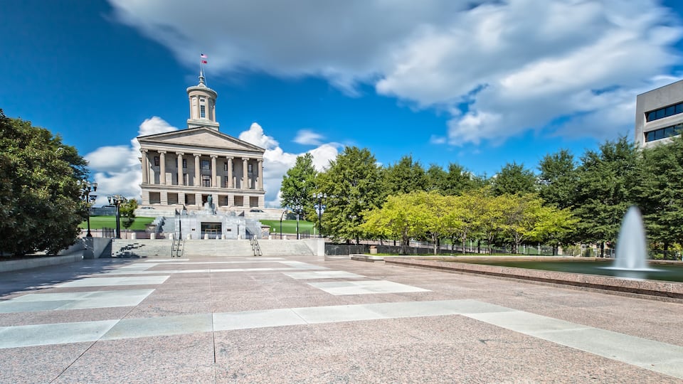 A neoclassical building with a central tower and flag, surrounded by a plaza with a fountain and trees.