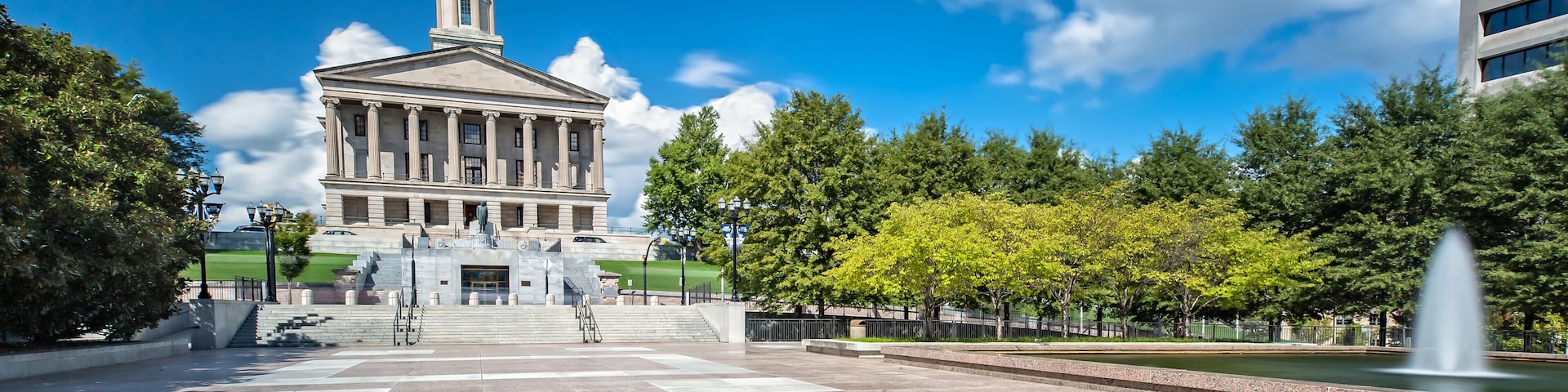 A neoclassical building with a central tower and flag, surrounded by a plaza with a fountain and trees.