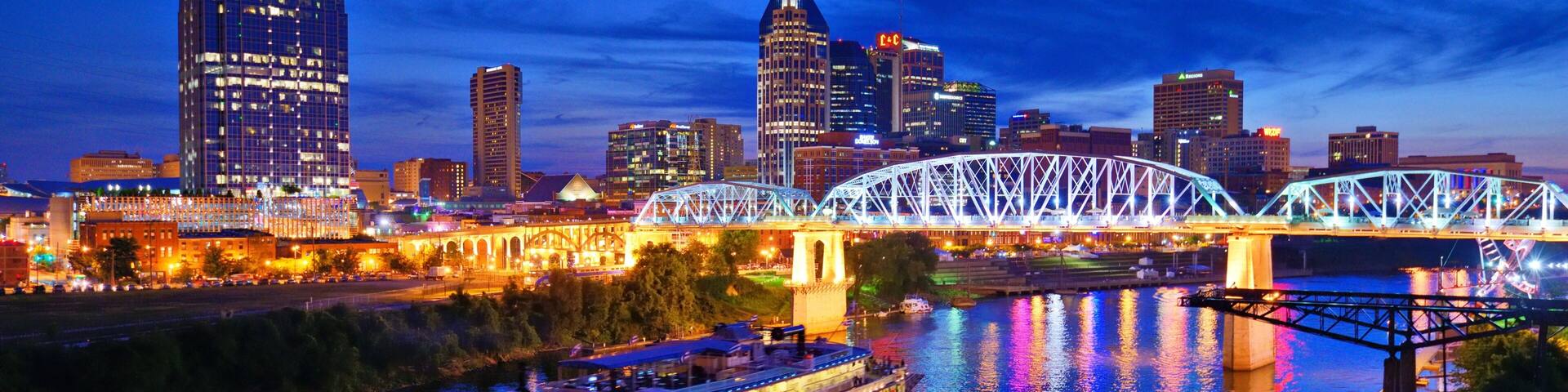 A river with a boat, a bridge, and a city skyline with illuminated buildings.