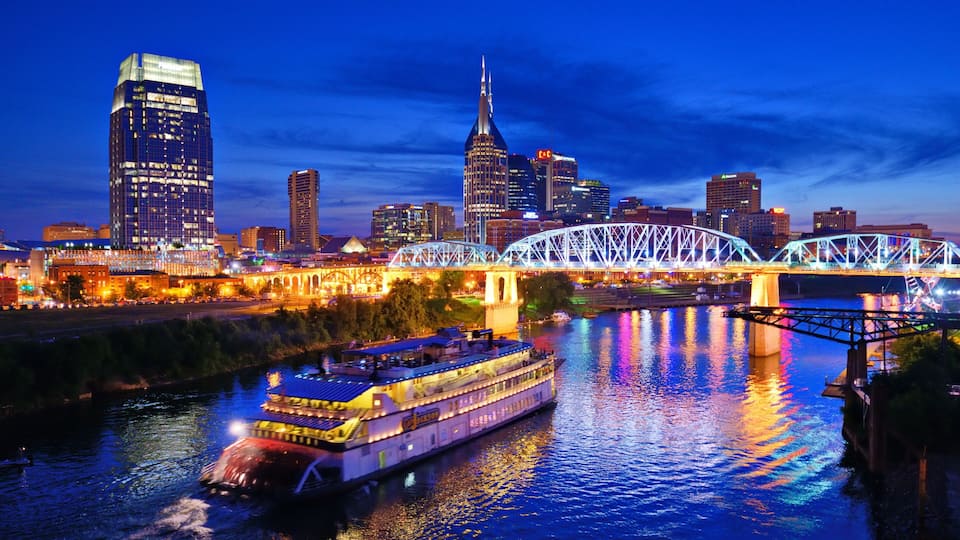 A river with a boat, a bridge, and a city skyline with illuminated buildings.