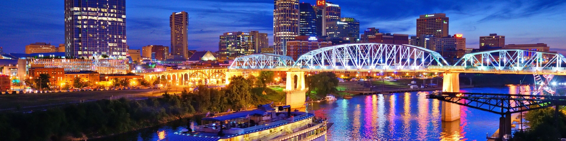 A river with a boat, a bridge, and a city skyline with illuminated buildings.