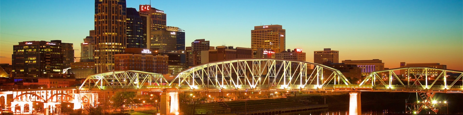 A city skyline with a prominent skyscraper and a brightly lit bridge over a river.