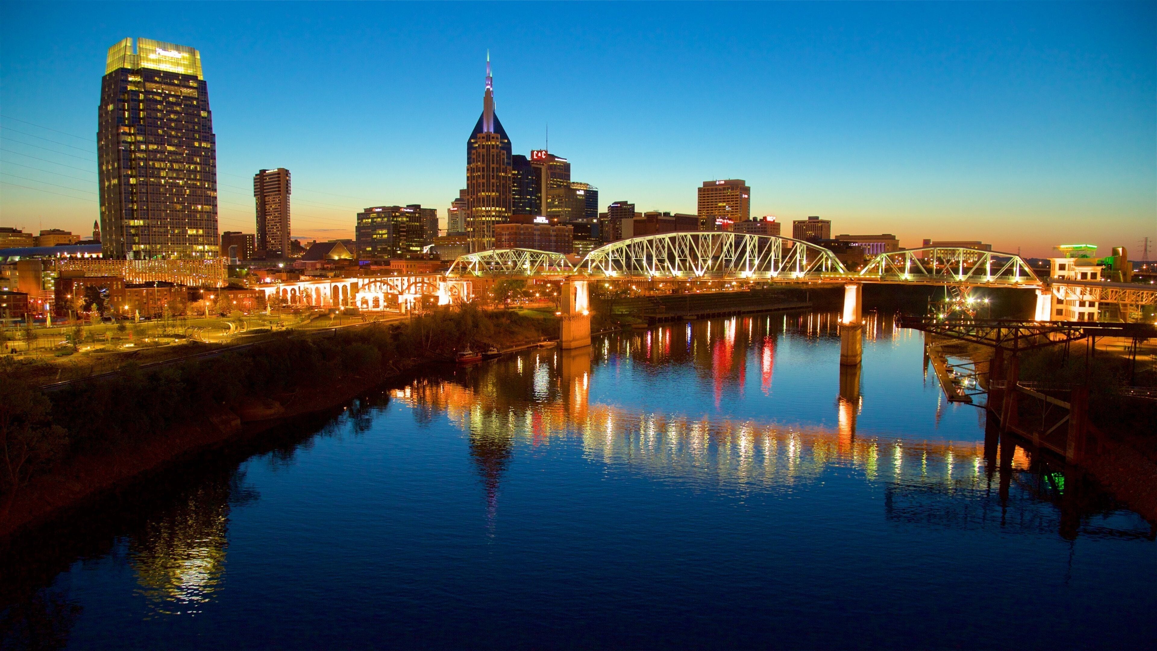 A city skyline at dusk with illuminated buildings and a bridge over a river reflecting the lights.