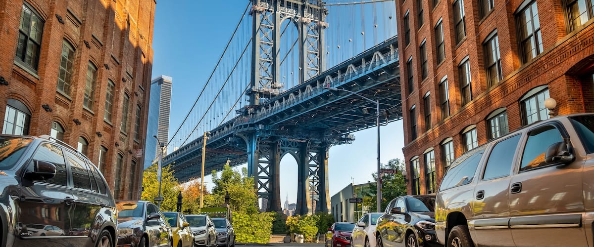 A street with parked cars and a large bridge in the background.