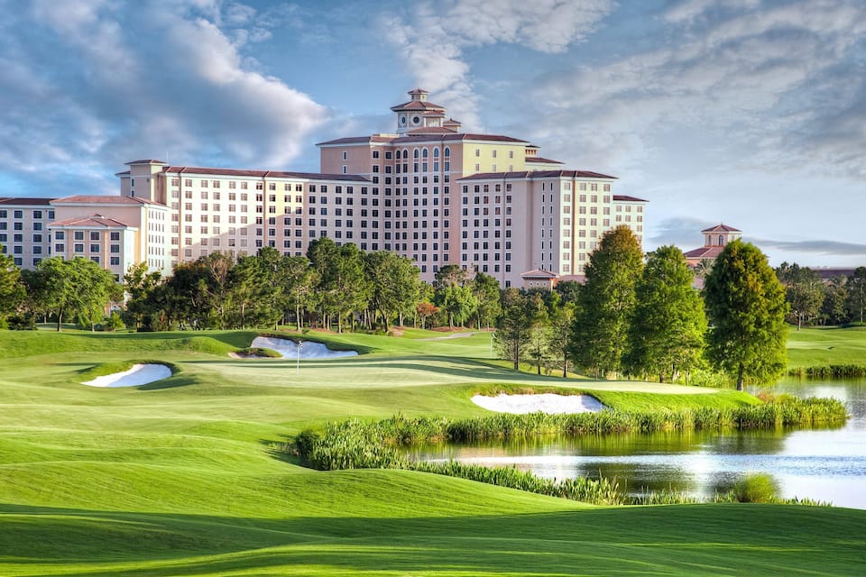 A golf course with bunkers, a pond, and a large hotel building in the background.